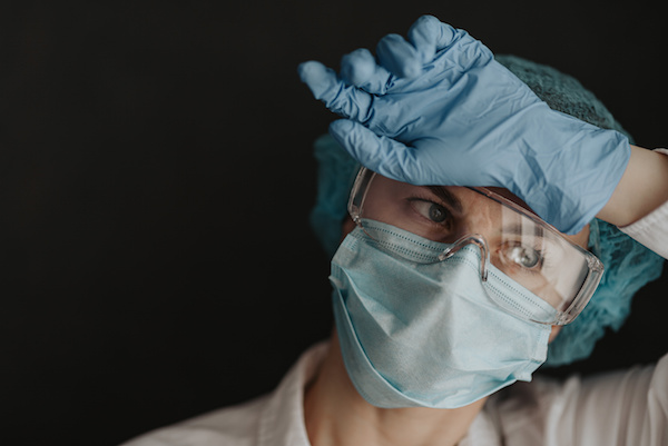 essential worker in protective mask and gloves rests her hand against her forehead